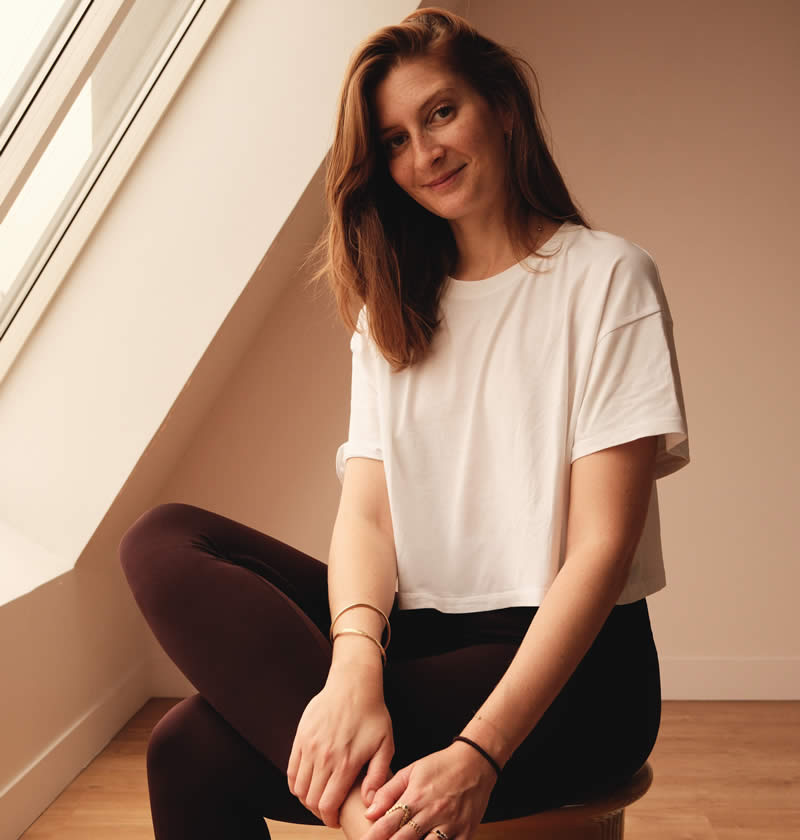 Oriane, professeure de Pilates postnatal, souriante, vêtue d’un t-shirt blanc et d’un pantalon de Pilates noir, regardant la caméra.