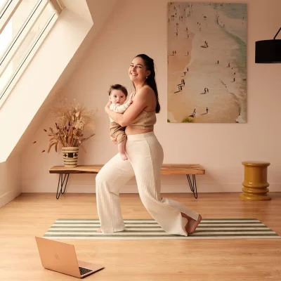 Maman tenant son bébé et pratiquant le Pilates postnatal devant son ordinateur.